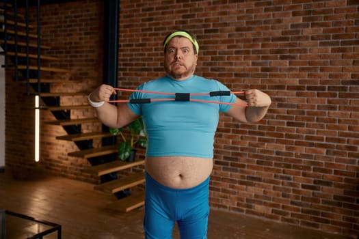 Funny chubby man in sportswear fooling around with fitness equipment during training at home. Fat sportsman crazy emotion