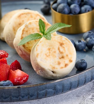 Round small fried cheesecakes on a blue plate with berries. Healthy breakfast