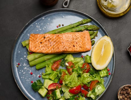Healthy lunch with grilled salmon with asparagus and fresh tomato and cucumber salad, a row of lemon slice. Food on a blue round plate, top view