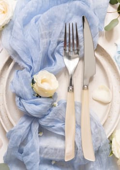 Wedding table place with fork and knife on plate, light blue tulle fabric, proposal ring and cream flowers, top view. Romantic pastel table decor