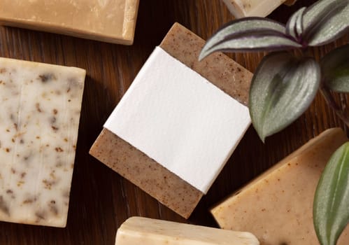 Beige and brown handmade soap bars with blank labels on wood near green plant top view, packaging mockup, copy space. Natural herbal products for face and body hygiene