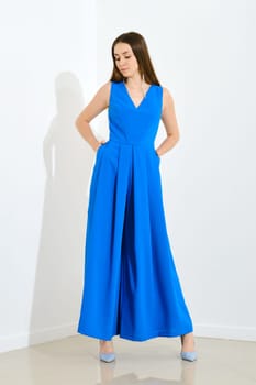Pretty stylish young woman dressed in blue long sleeveless dress posing over bright wall