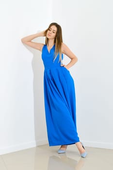 Dreamy young woman in long blue sleeveless dress leaning to white wall