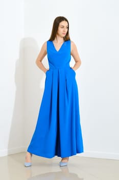 Full length portrait of stylish trendy woman in blue long sleeveless dress holding her hands in pockets