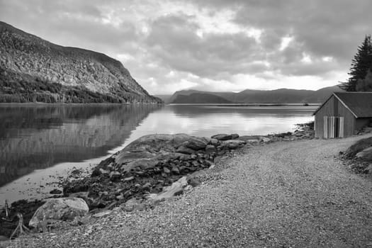 Norwegian landscape in black and white. View over a small harbor to the fjord with a cloudy sky behind the mountain. Scandinavian nature