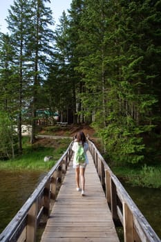 A girl with a backpack on her back walks along a wooden bridge in a quiet forest under a clear sky