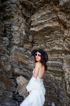 A woman in a white dress and black hat stands against a rugged rock formation