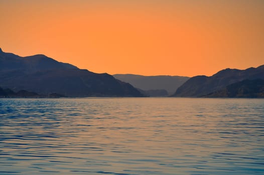 Norwegian fjord landscape at dusk. View of snow-capped mountains with rugged vegetation. Crystal clear water in the foreground. Landscape from Scandinavia