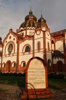 At the Synagogue of Subotica at golden hour, the memorial stone, commemorative stone in the foreground and the illuminated facade of the Synagogue in the background, Subotica, Serbia 2022