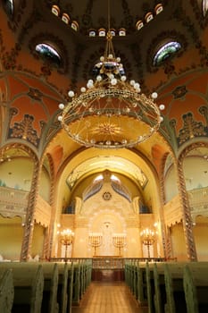 Inside the Synagogue of Subotica, a big golden chandelier is hanging from the colorful ceiling over the the benches and the aisle leading to the altar, Subotica, Serbia 2022