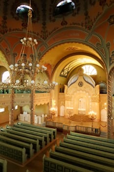 Inside the Synagogue of Subotica, a big golden chandelier is hanging from the ceiling over the benches and the aisle leading to the illuminated altar, Subotica, Serbia 2022