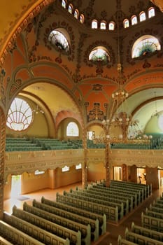 Inside the Synagogue of Subotica, a big golden chandelier is hanging from the painted ceiling over the benches and the aisle, light is shining through stained glass windows in the background, Subotica, Serbia 2022