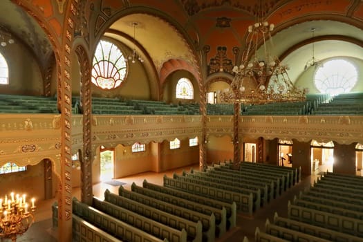 Inside the Synagogue of Subotica, a big golden chandelier is hanging from the ceiling over the benches and the aisle, light is shining through stained glass windows in the background, Subotica, Serbia 2022