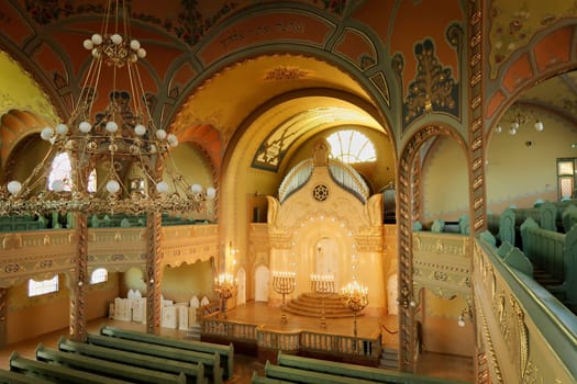 Inside the Synagogue of Subotica, a big golden chandelier is hanging from the ceiling over the benches and the aisle leading to the altar illuminated by candles, Subotica, Serbia 2022