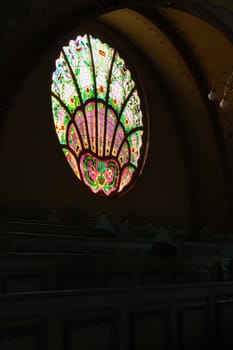 Stained glass window in different shades of purple next to wooden benches and a chandelier inside the Synagogue of Subotica, second floor, Subotica, Serbia 2022