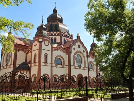 View between trees onto the Synagogue of Subotica behind a black metal fence with elaborate details on a sunny day, Subotica, Serbia 2022