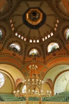 Inside the Synagogue of Subotica, the big golden chandelier is hanging from the painted ceiling, vault, dome, light is shining through stained glass windows, Subotica, Serbia 2022