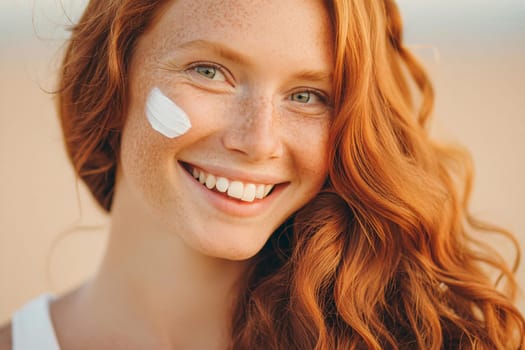Close-up face of happy long-haired smiling young woman with red hair, freckles and pigmentation on her face with a smear of sunscreen on her skin. Protects the skin from pigmentation and dark spots in the sun
