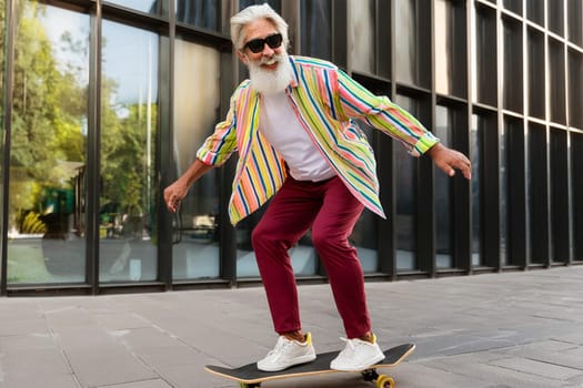 Elderly gray-haired man with beard in colored clothes enjoying skateboarding ride in city.