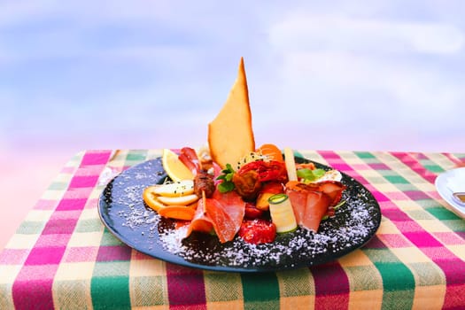 Traditional Italian food. Close-up of mixed Mediterranean appetizers or antipasto and tapas with ham vegetables and crab on black plate on table over bright background. Space.