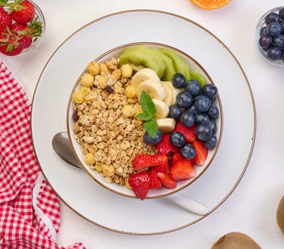 Granola with strawberries, kiwi, banana and blueberries in a round plate on a white table. Healthy and tasty food, top view
