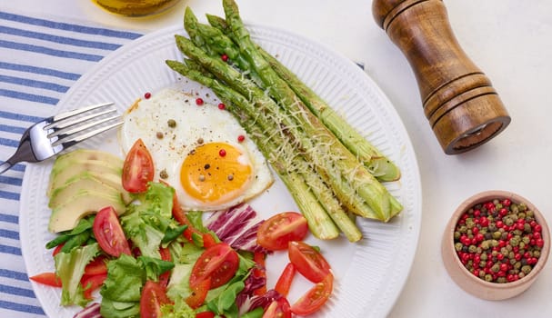 Round plate with cooked asparagus, fried egg, avocado and fresh vegetable salad on the table, top view