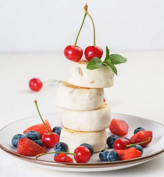 Stack of four cheesecakes, decorated with two cherries and a sprig of mint, on a plate with berries, blueberries, strawberries and cherries. Background white