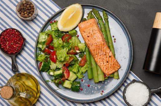Healthy lunch with grilled salmon with asparagus and fresh tomato and cucumber salad, a row of lemon slice. Food on a blue round plate, top view