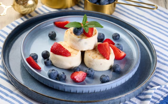 Round small fried cheesecakes on a blue plate with berries. Healthy breakfast, top view
