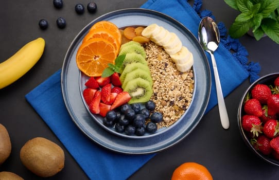 Granola with strawberries, kiwi, banana and blueberries in a round plate on a black table. Healthy and tasty food, top view