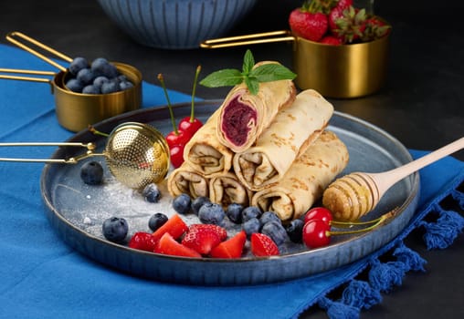 Crepes stacked in a pile, one of which is cut and filled with berry filling. The plate is also decorated with blueberries, strawberries, and cherries, a honey dipper with honey, a sprig of mint