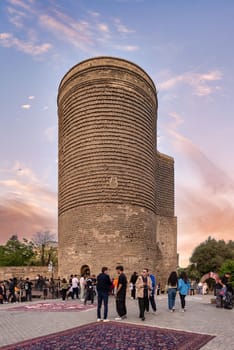 Baku, Azerbaijan - May 4, 2024: Tourists gather at the base of the Maiden Tower under a beautiful sunset sky. The ancient tower stands tall, its brickwork illuminated by the warm evening light