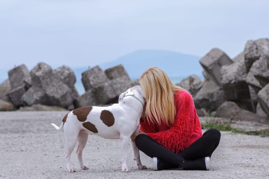 Sitting on the ground a blonde with a black wedge and a red blouse kisses a pit bull on the ear