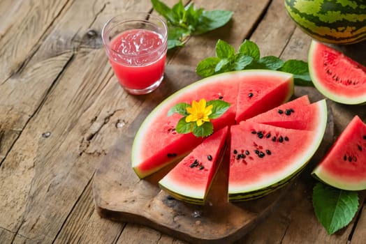 Close-up of fresh watermelon slices with a glass of watermelon juice on a rustic wooden table, garnished with mint leaves and a yellow flower.