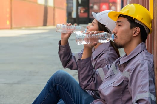 Male and female engineers in uniform take a break to drink water from plastic bottle after finish routine work of containers loading at carto shipping yard
