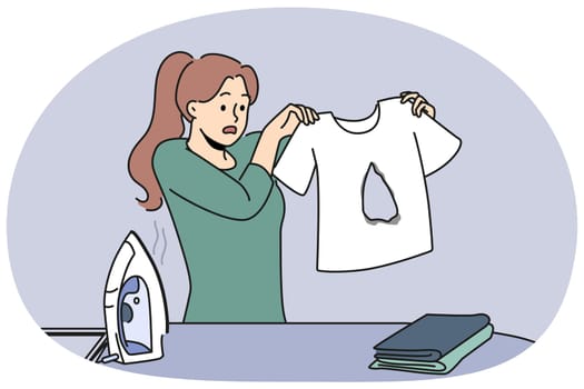 Clumsy housewife ironing clothes burnt hole in t-shirt and dont know how to fix mistake. Woman stands near ironing board with clothes and iron and looks at damaged item in confusion.