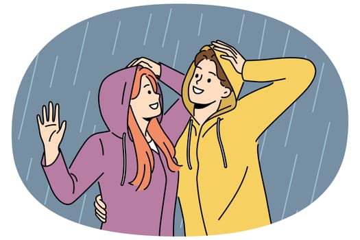 Happy couple stands in rain in waterproof hoods and hugs rejoicing at cloudy weather and first autumn downpour. Cheerful man and woman walking in rain during date or having romantic conversation