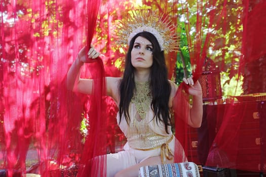 A woman dressed as a goddess looks through red curtains