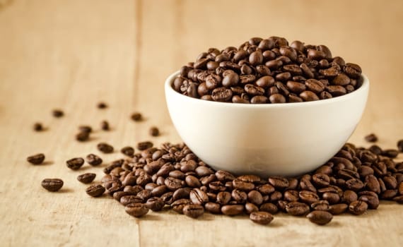 A white bowl filled with coffee beans, with more beans scattered around, placed on a wooden surface.