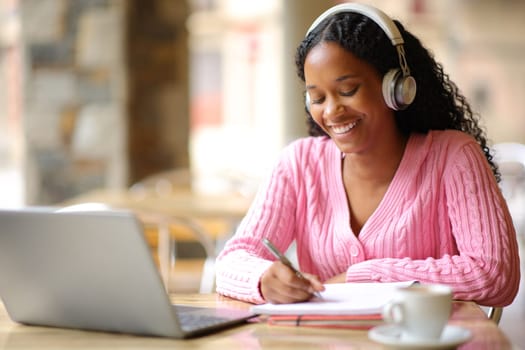 Happy black student taking notes e-learning with laptop and headphone in a bar