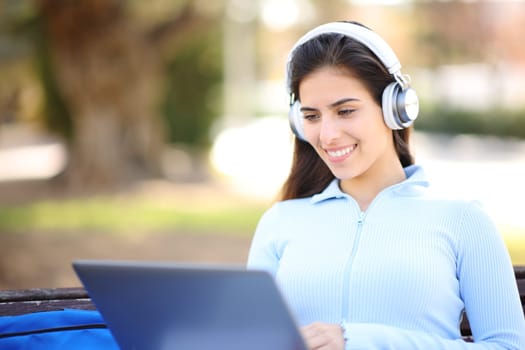 Happy student e-learning in a park using headphone and laptop sitting in a bench