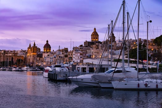 Birgu city, part of the Three Cities of Malta. Night marina glows under the night sky, yacht masts silhouetted against twinkling lights reflecting on the tranquil waters. Travel destination.