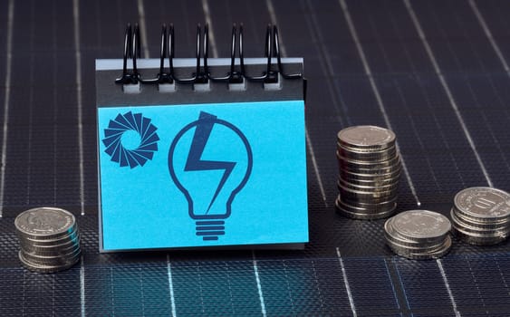 A stack of coins and a notepad with a drawn light bulb stand on the surface of a solar panel, alternative renewable energy concept