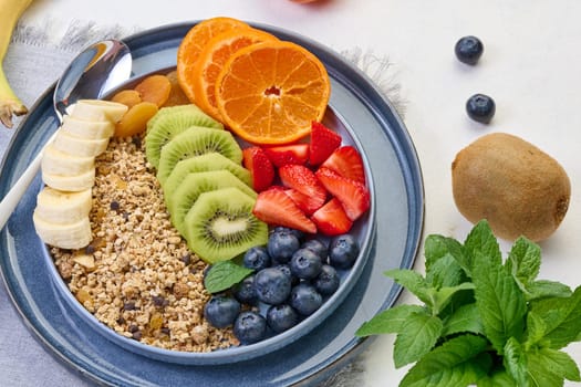 Granola with strawberries, kiwi, banana and blueberries in a round plate on a white table. Healthy and delicious food
