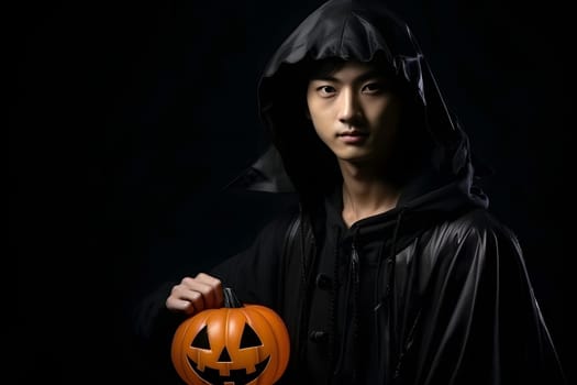 A young Asian man is standing in a dark setting, wearing a black hooded cloak and holding a carved Halloween pumpkin