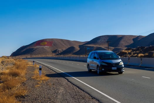 Car is driving along the road through natural mountain landscape with the flag of Kyrgyzstan is drawn on a mountain at sunny day in Kok Moynok Eki, Kyrgyzstan - December 12, 2024