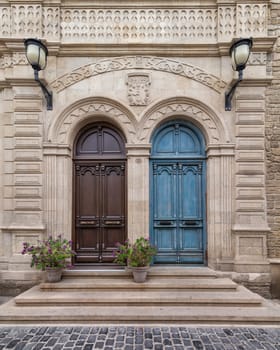 Showing a blue and a brown wooden doors side by side, this picture highlights the contrast between two different architectural styles. The building is decorated with street lights and plants