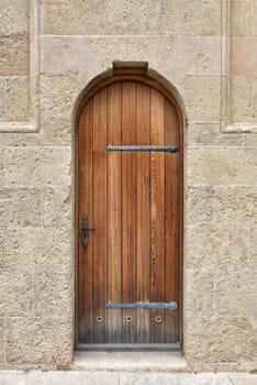 A close-up image of a weathered, arched wooden door set within a stone wall. The door is made of dark brown wood with horizontal planks and has a metal handle and two large metal hinges