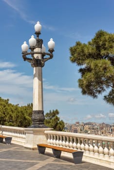 A white stone lamp post with ornate black metal details, stands tall on a stone walkway overlooking a city with buildings and trees in the distance