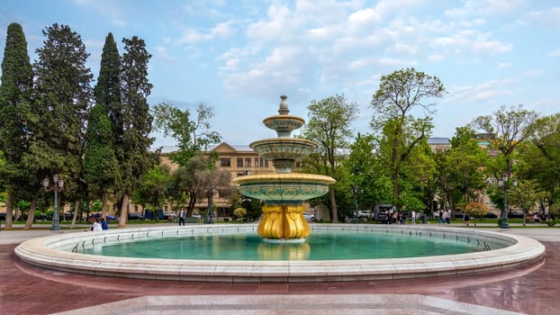 Baku, Azerbaijan - May 5, 2024: In Bakus Sahil Bagi park, a golden based fountain with blue and white tiles sits in a circular pool, surrounded by lush trees and a walkway